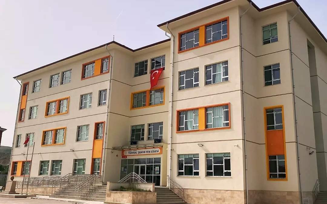 Şeker Fen Lisesi binasının krem ve turuncu cepheli ön görünümü, önünde banklar